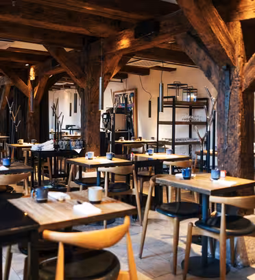 Mysig restauranginteriör med träbjälkar och bord. Sittarrangemanget är snyggt organiserat med naturligt ljus. Minimalistisk inredning och mjuk belysning skapar en varm atmosfär.