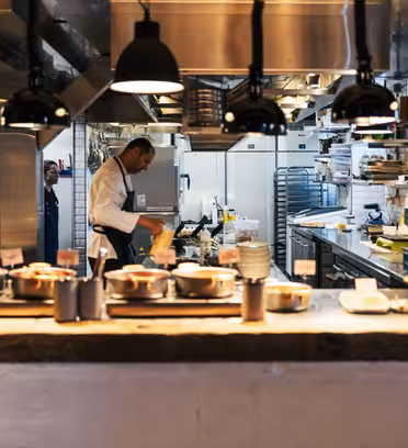 En kock i vit rock och förkläde lagar mat i ett hektiskt, modernt restaurangkök. Rätterna är snyggt ordnade på en bänk och industriköksutrustning syns i bakgrunden. Taklampor lyser upp arbetsytan.