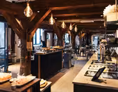 En rymlig bufférestaurang i rustik stil med synliga träbjälkar och varm belysning. Borden är dukade med en mängd olika rätter, och stora fönster tillåter naturligt ljus att fylla utrymmet.