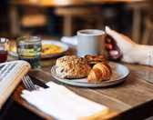 En person som håller i en mugg sitter vid ett träbord med en tallrik med bakverk, inklusive en croissant och en frörulle. I närheten finns en tidning, ett glas juice och ett annat glas. Bakgrunden är mjukt suddig.
