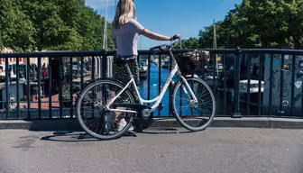 En person med långt hår står på en bro med utsikt över en flod och håller i en vit cykel med en korg. Floden är kantad av båtar och omgiven av grönskande träd under en klarblå himmel.