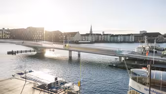 En solbelyst scen vid vattnet med en modern gångbro som sträcker sig över en lugn flod. Byggnader kantar vattnet och ett par båtar ligger i anslutning till. Himlen är klar och solen kastar reflektioner på vattnet.