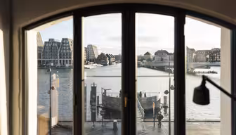 En vy genom ett välvt fönster visar en scen vid vattnet med moderna och traditionella byggnader. En lugn flod speglar den molniga himlen, medan en strandpromenad full av människor väcker den lugna stadsbilden till liv.