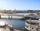 En naturskön utsikt över en hamn i Köpenhamn, med en gångbro med människor som går och cyklar. Båtar lägger till i närheten och historiska byggnader kantar vattnet under en klarblå himmel.