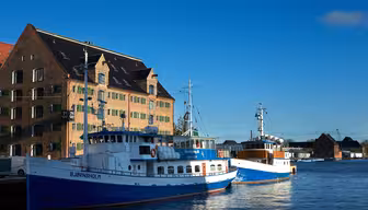 Två blåvita båtar ligger förtöjda nära en historisk tegelbyggnad vid vattnet. Himlen är klar och ljus och kastar skarpa skuggor på båtarna och byggnaden. Scenen är lugn med minimal aktivitet runt omkring.