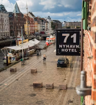 Utsikt från en tegelbyggnad som visar en kullerstensgata bredvid en kanal med båtar förtöjda. På en skylt står det "71 Nyhavn Hotel." Människor går längs strandpromenaden under en delvis molnig himmel, med färgglada historiska byggnader i bakgrunden.