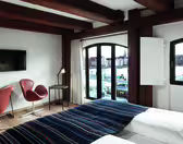 Ett mysigt hotellrum med träbjälkar har en säng med en randig filt, två bruna stolar, en tv och ett skrivbord. Stora välvda fönster erbjuder utsikt över en strand och byggnader över vattnet.
