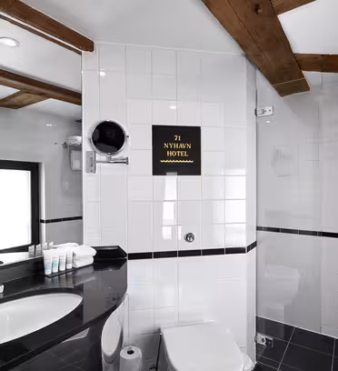 Modernt badrum med vitt kakel och träbjälkar. Har en svart diskbänk med toalettartiklar, en rund spegel och en vägg med en liten plakett där det står "71 NYHAVN HOTEL". Handdukshängare rymmer vita handdukar; en duschkabin är delvis synlig.