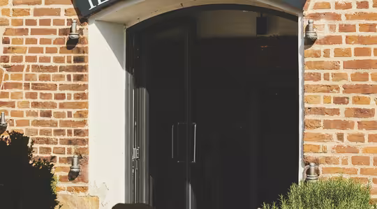 Entré till restaurangen "Il Rosmarino" med öppen dörr i en tegelbyggnad. Scenen har utomhusbord dukade med glas och korgstolar, under stora paraplyer. Frodiga gröna växter ger en touch av naturen.