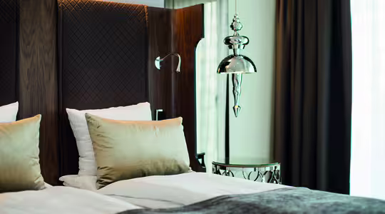 Ett modernt hotellrum har en snyggt bäddad säng med vita sängkläder, beiga kuddar och en brun quiltad sänggavel. En pendellampa i silver och ett litet runt sängbord med reflekterande yta finns i närheten. Mörka gardiner ramar in ett fönster.